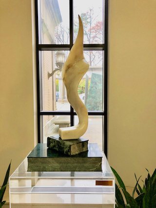 Knowledge is Eternal marble statue on green marble and lucite pedastal placed by a window; the color is pale yellow and the shape is pointed at the top with soft curves in the middle and at the bottom